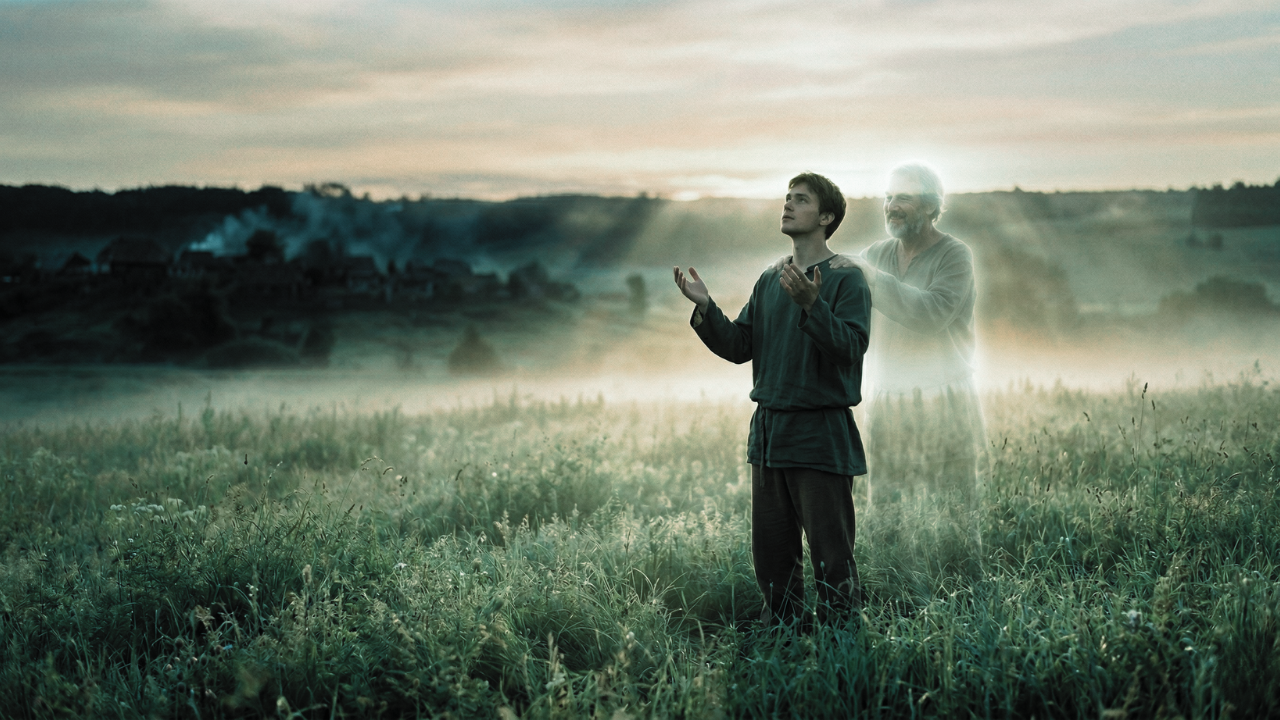 A lone man stands in a vast golden field at dawn, arms raised toward a breaking sky